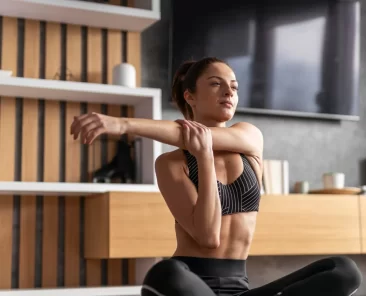 Mulher faz alongamento de ombro sentada no chão, em ambiente moderno e iluminado, vestindo roupa esportiva.