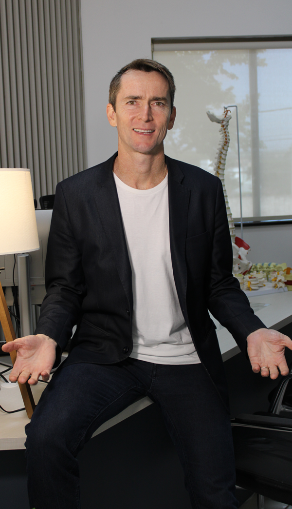 Imagem do Dr. Alexandre Jaccard, ortopedista Avaré, homem branco, sorrindo, vestindo camiseta branca e blazer preto, em consultório médico.