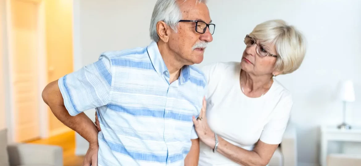 Um homem idoso sente dor na coluna e é amparado pela sua esposa.