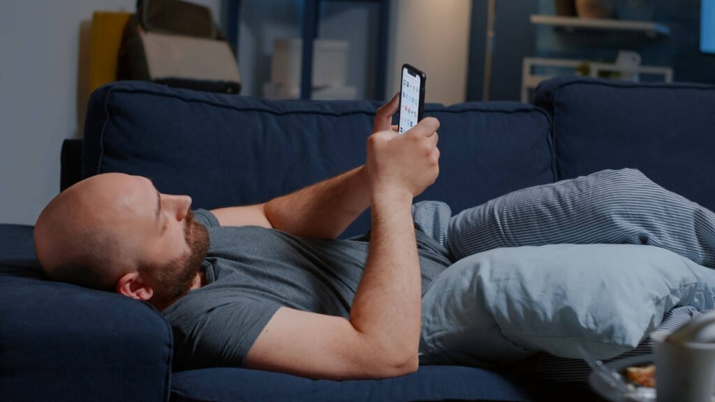 um homem sedentário mexe no celular deitado no sofá.